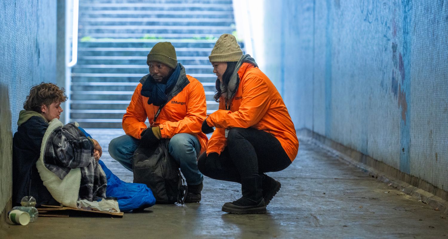 Two St Mungo's outreach workers in orange jackets crouch next to a man sleeping rough in an underpass. 