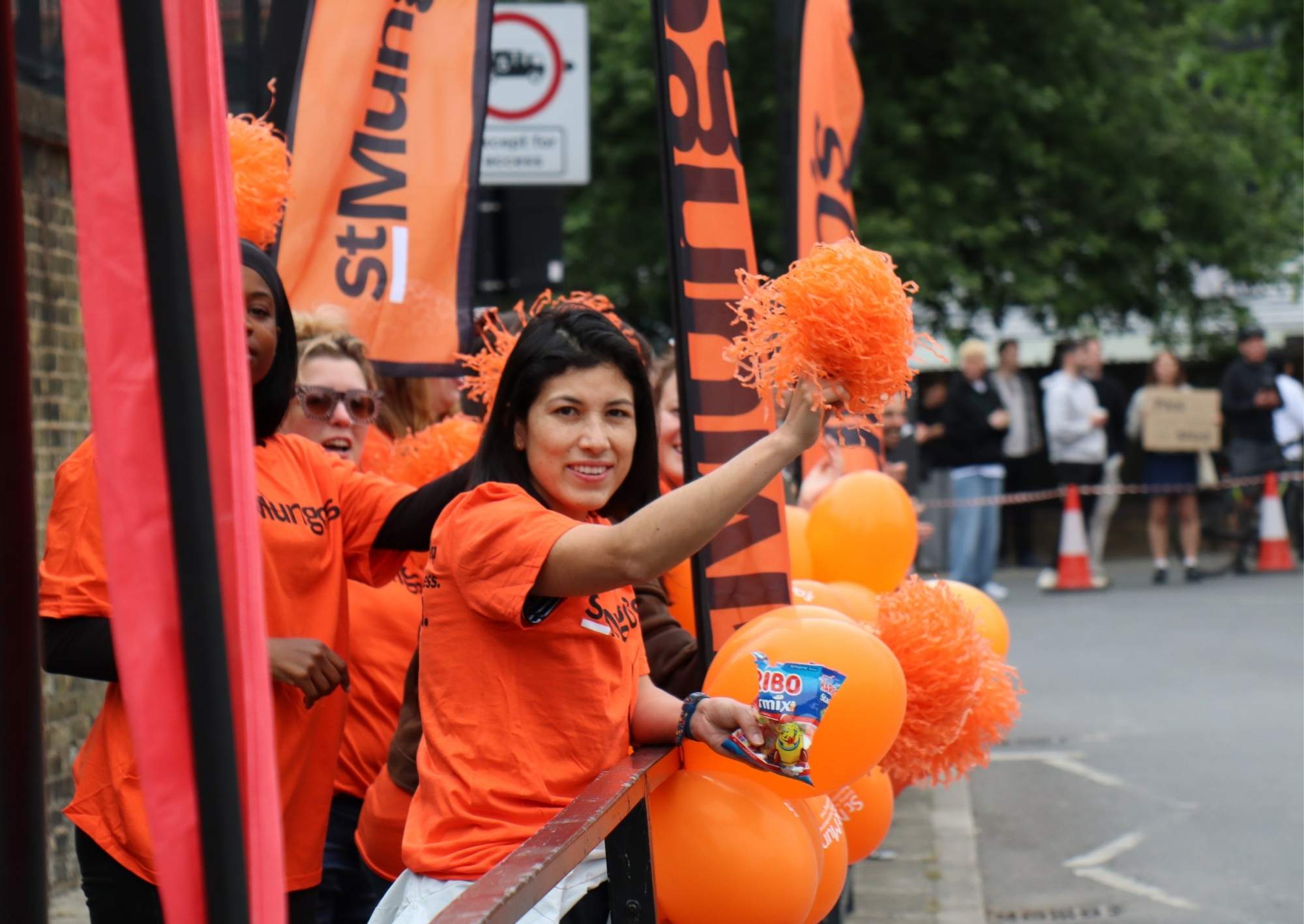 A team of people wearing the orange St Mungo's tee shirt cheering with balloons and pom poms.