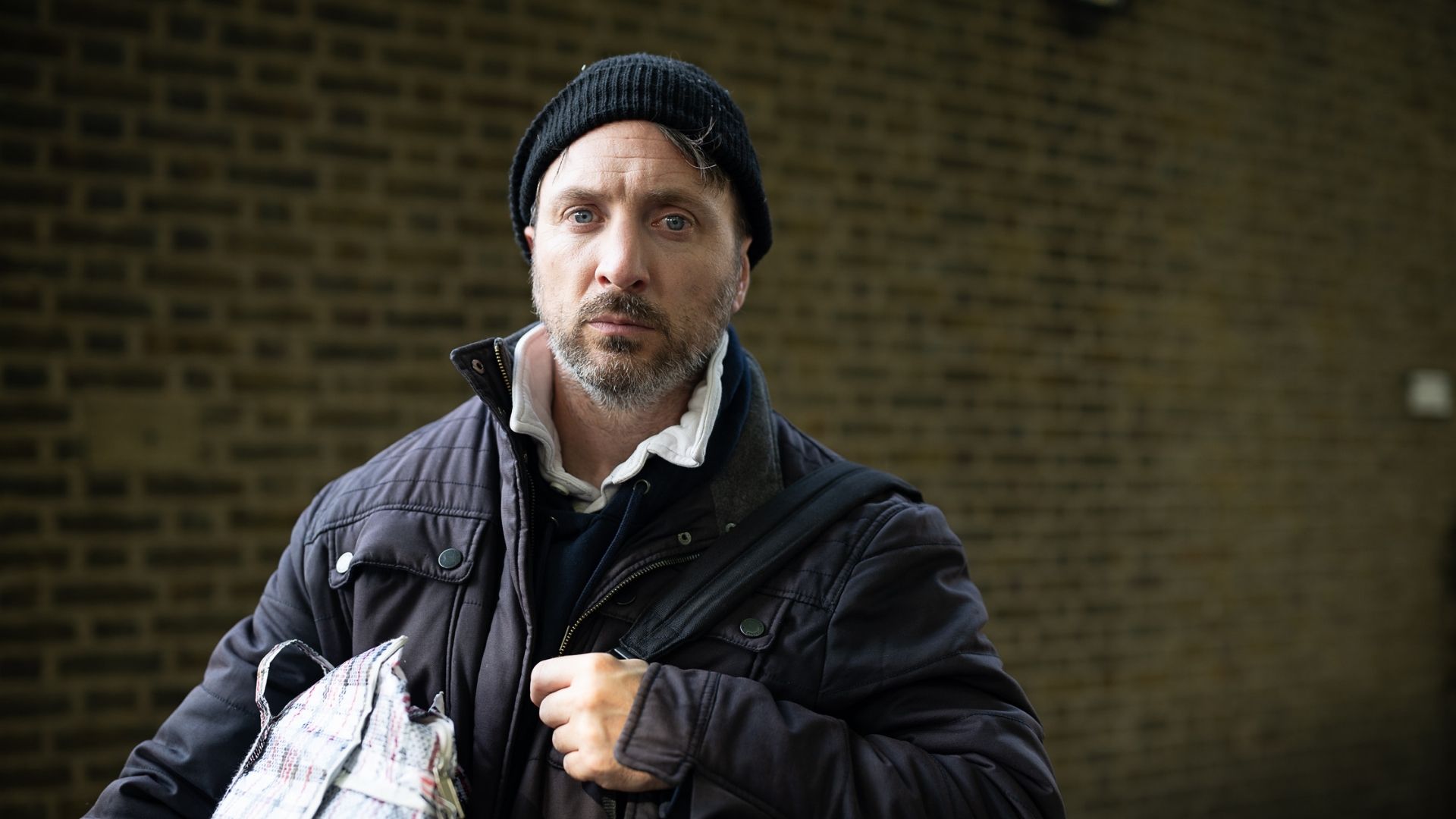 A man looking into the camera, carrying a bag, wearing a jacket and a winter hat.