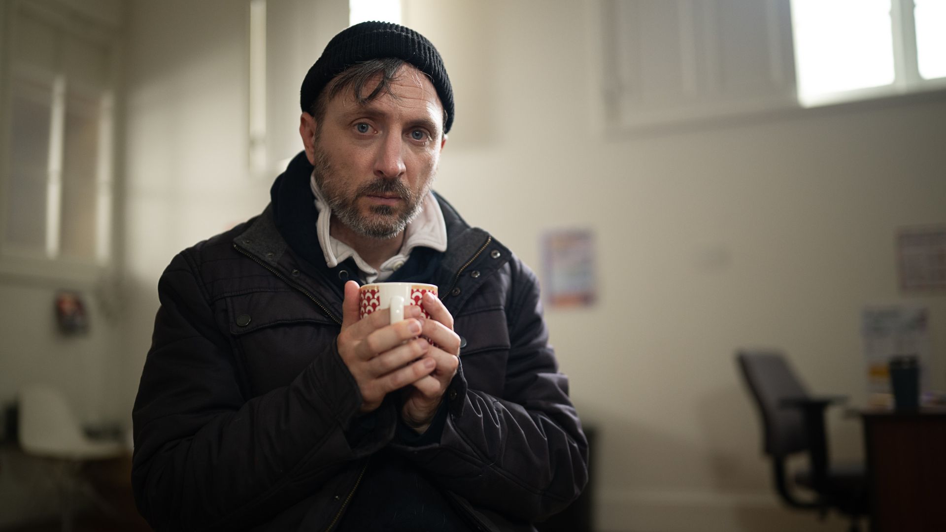 A man looking into the camera, sitting with a mug in hand, wearing a jacket and a winter hat.