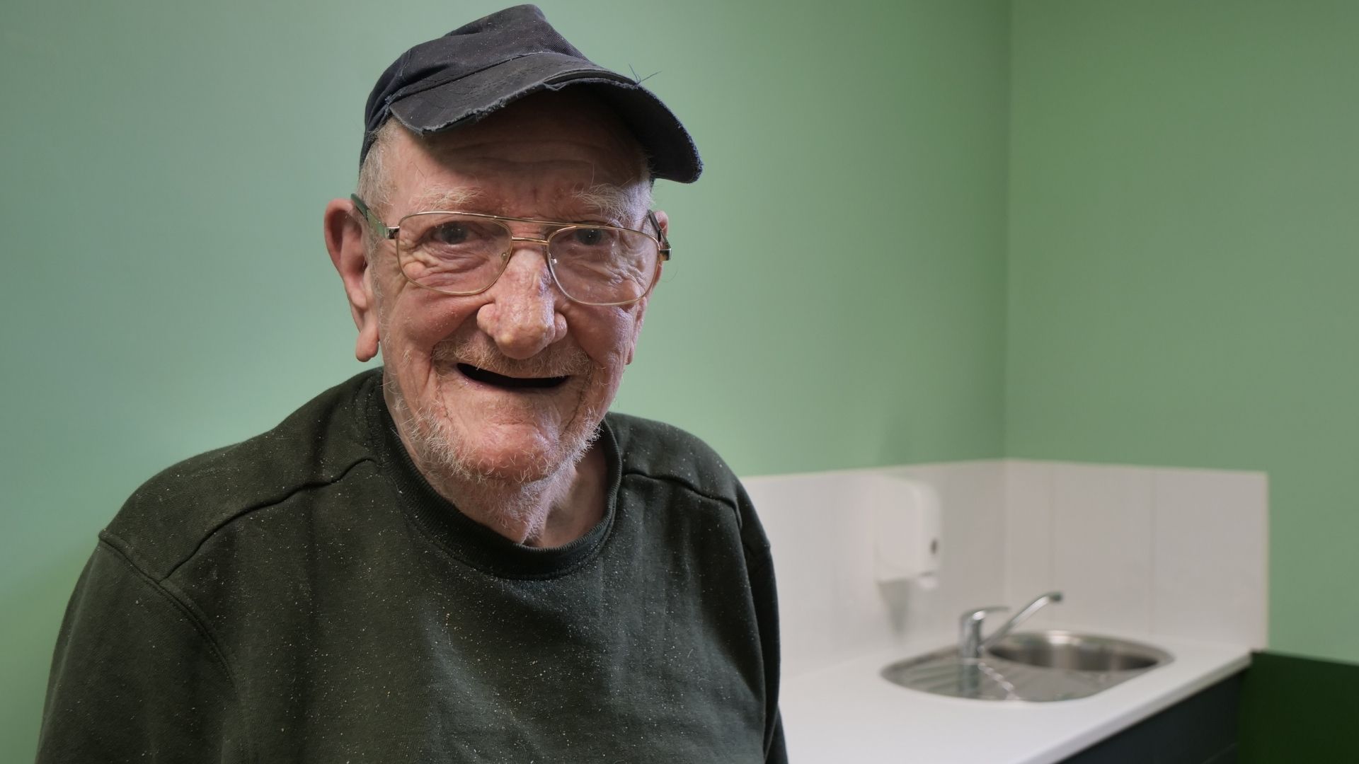 A man looking into the camera, wearing a face cap and glasses. A green wall showing in the background and a sink by the corner.