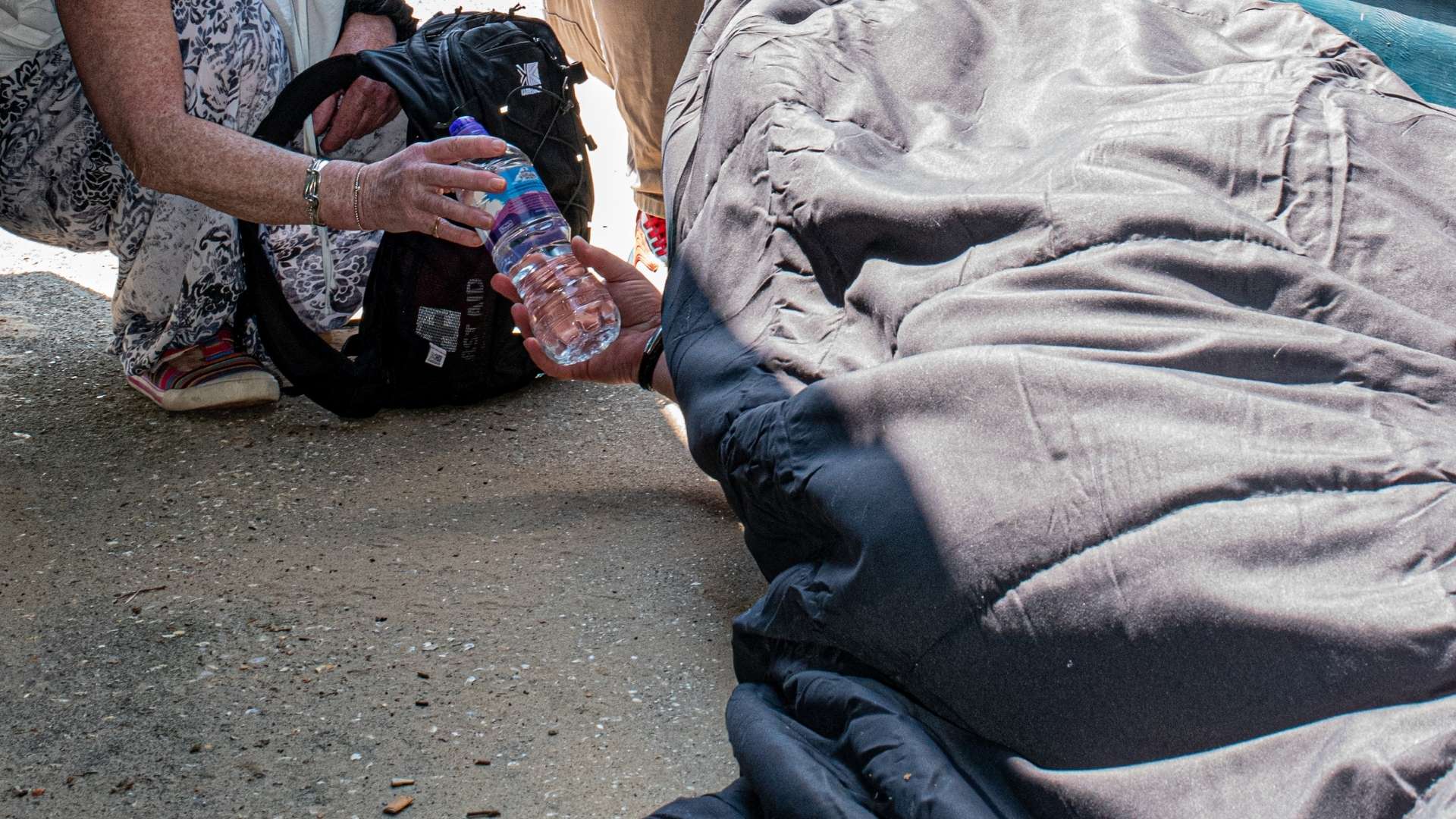 A rough sleeper in the hot sun, being offered a drink.