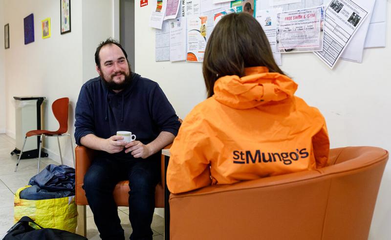Two people have a conversation in a St Mungo's centre. One is holding a mug, the other is wearing an orange St Mungo's jacket with the logo on it